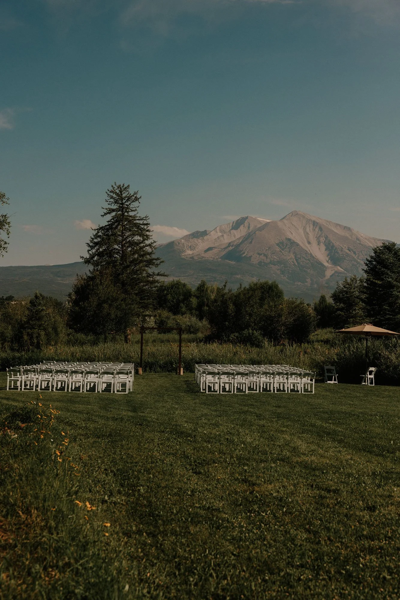 Outdoor wedding setup with rows of white chairs on a grassy lawn, with a mountain and trees in the background.