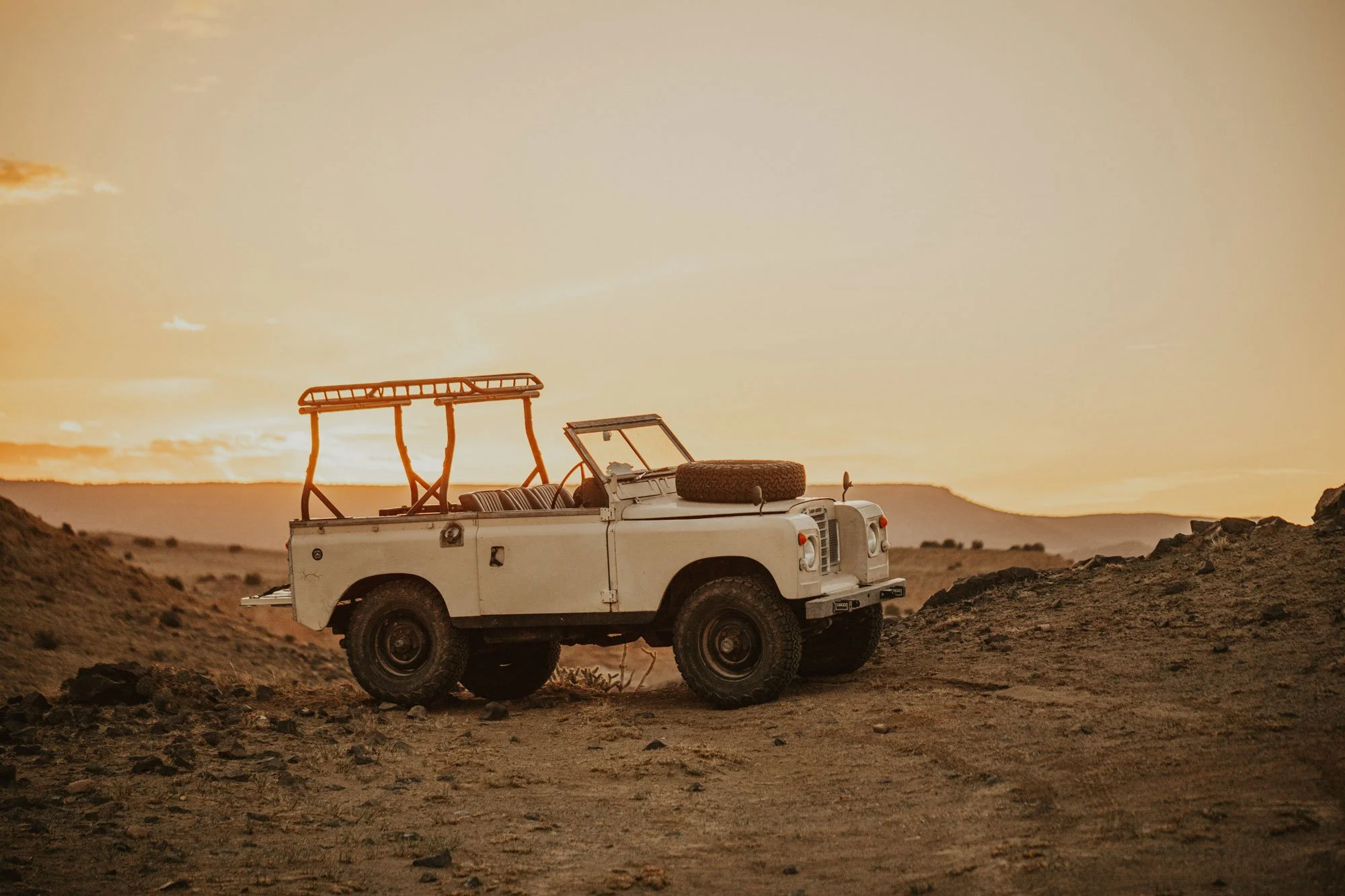 A white off-road vehicle with a spare tire on the hood, situated in a desert landscape during sunset with an orange sky and distant hills.