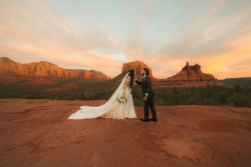 A bride and groom standing on a red rock formation during sunset with a scenic desert mountain landscape in Arizona. 
