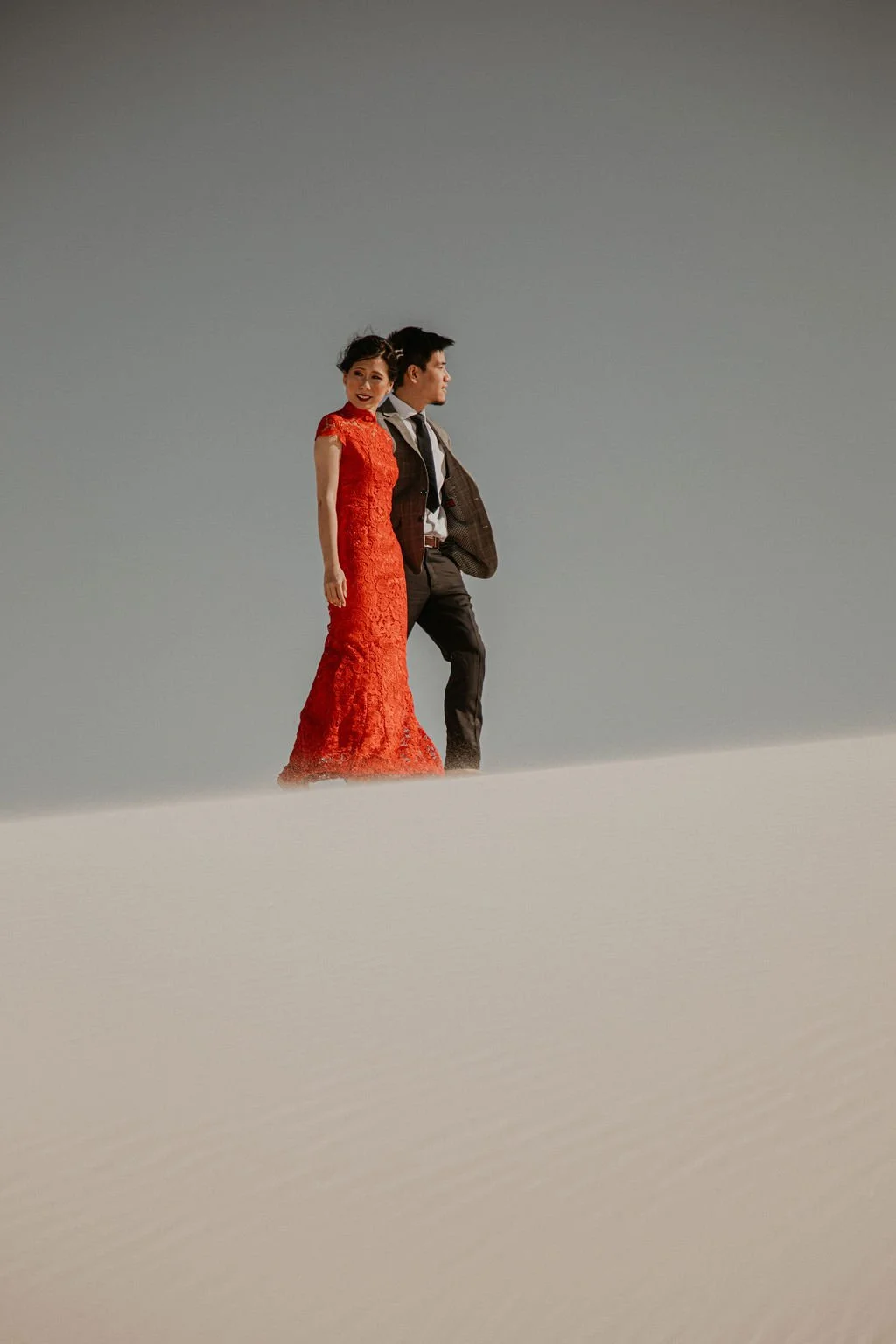 A woman in a red lace dress and a man in a suit standing on a barren, snowy landscape against a gray sky.