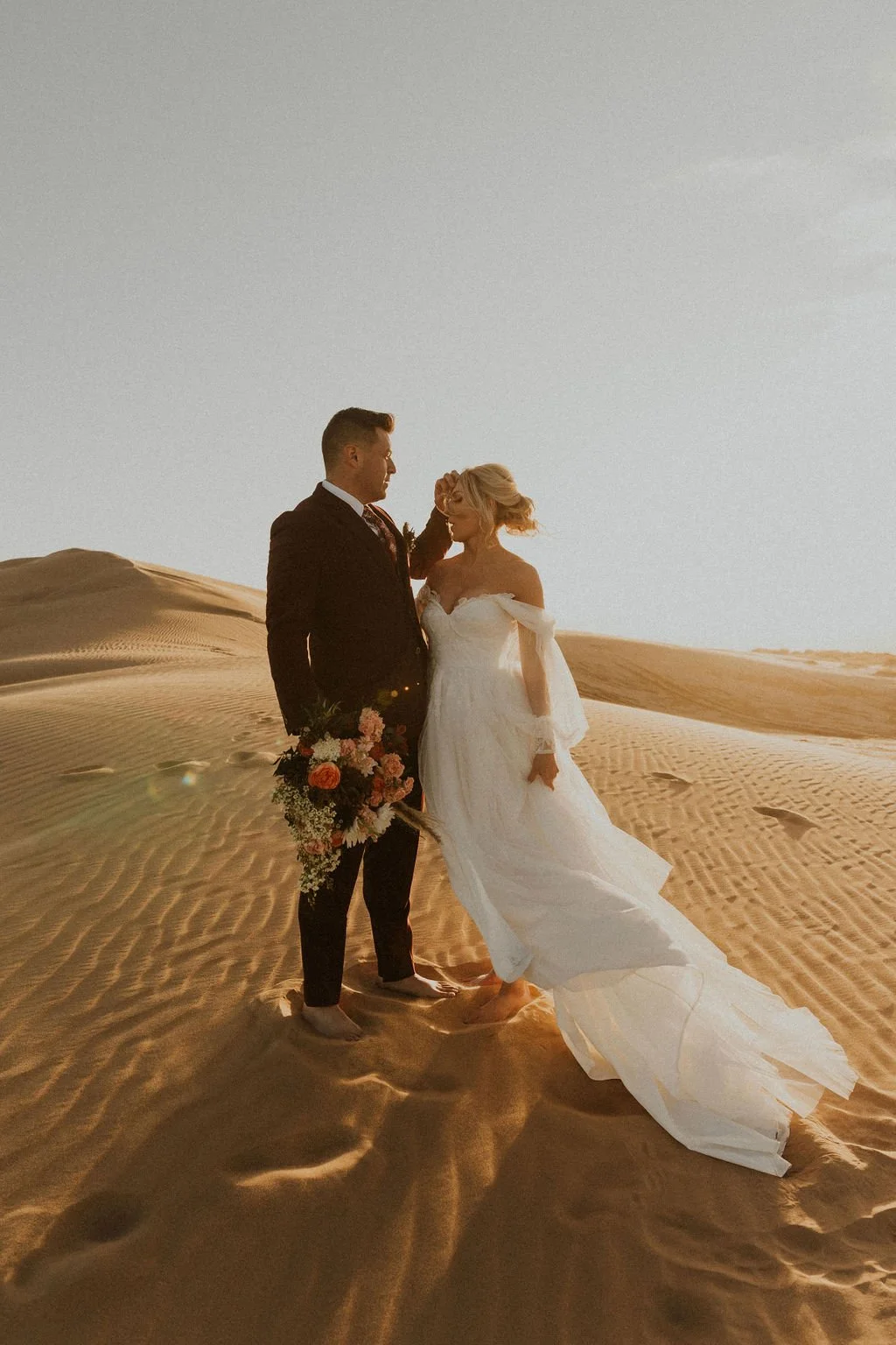 A bride and groom standing barefoot on sand dunes during sunset, with the groom holding a bouquet of flowers and the bride gently touching her face.