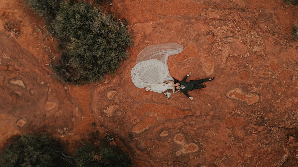 A bride and groom lying on the ground in a desert landscape with red rocks and sparse bushes, holding hands and looking up.