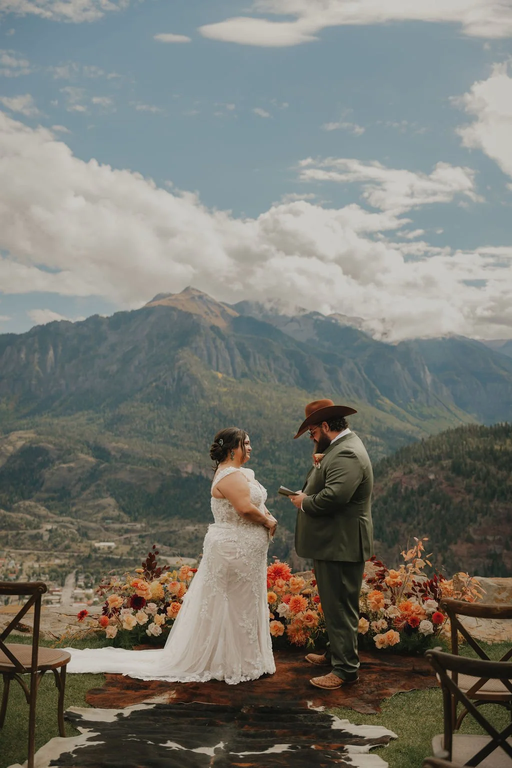 A bride and groom stand facing each other during a wedding ceremony outdoors with mountains and a partly cloudy sky in the background. The bride wears a white lace wedding dress, and the groom wears a gray suit, cowboy hat, and glasses. They are surr