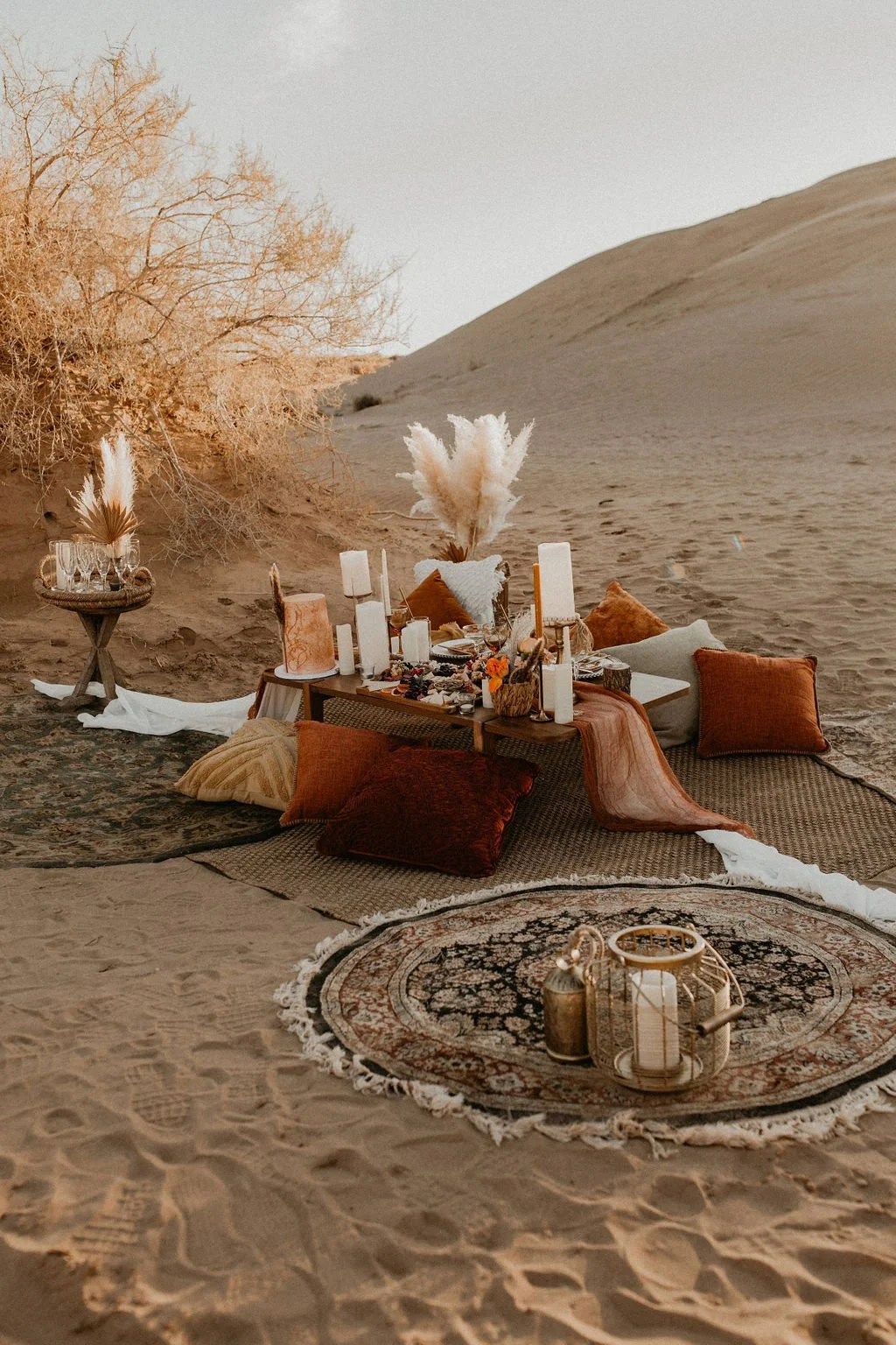 A cozy outdoor desert picnic setup with cushions, candles, and decorative items on rugs, set against sand dunes and a tree.