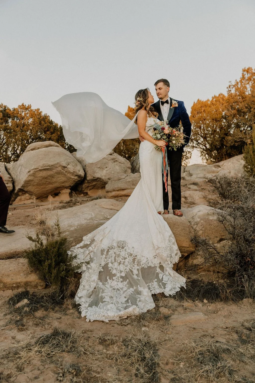 A bride and groom standing close together outdoors on rocky terrain during sunset. The bride wears a lace wedding gown with a long train and a flowing veil. The groom is in a tuxedo with a boutonniere. The bride holds a bouquet of flowers, and they a