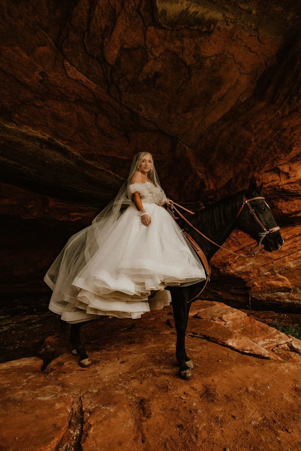 A woman in a white wedding dress sitting on a black horse inside a red rock cave