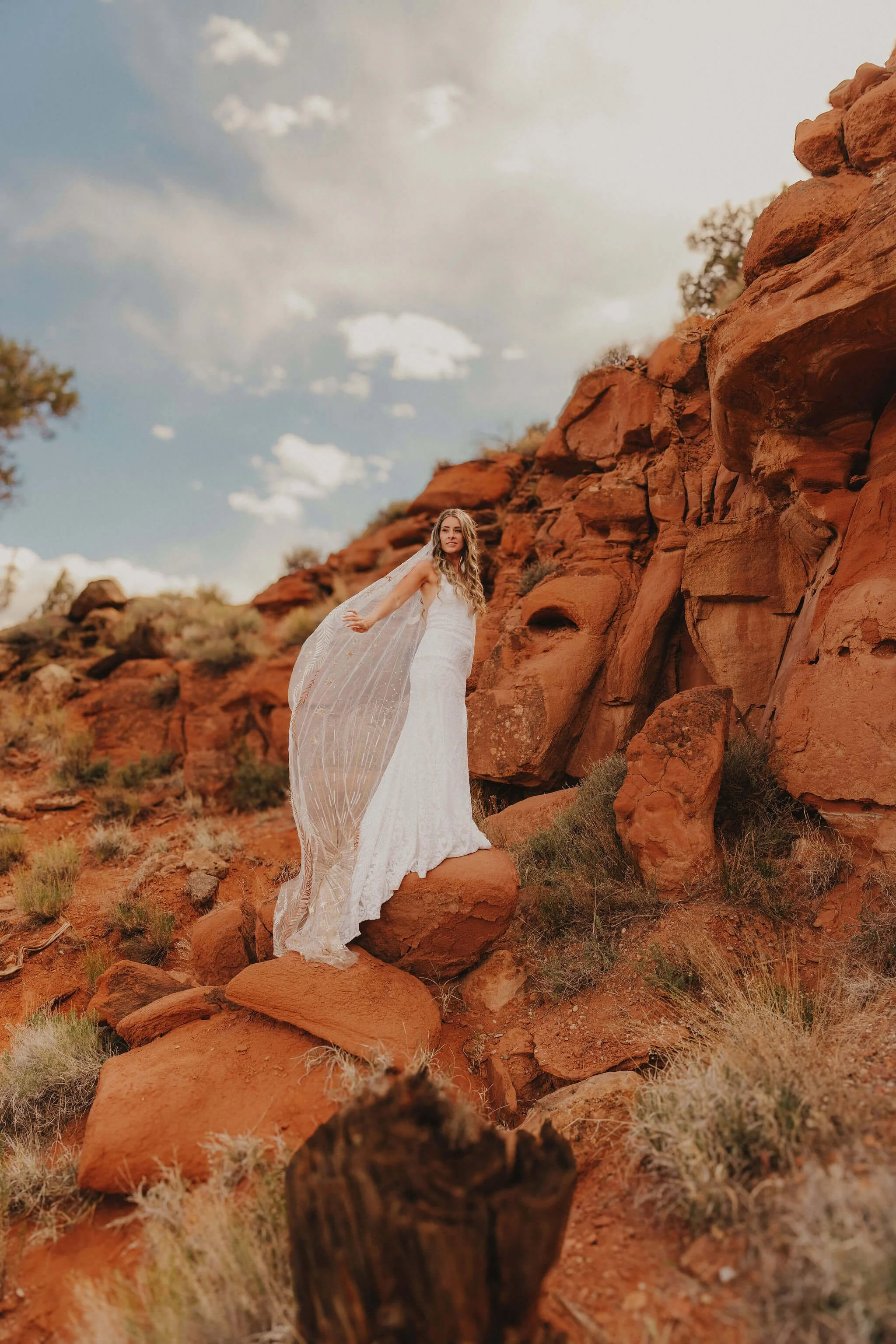 A woman in a white wedding dress standing on a large rock in a desert landscape with red rocks and bushes, under a sky with scattered clouds.