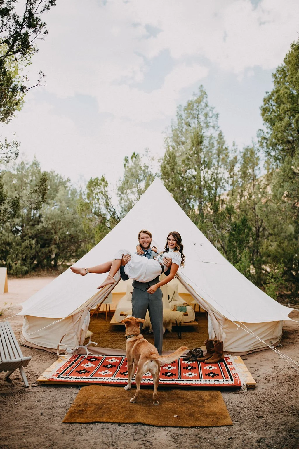 A couple is standing in front of a white tipi tent outdoors, with the man holding a woman in his arms. There's a dog standing on a colorful rug in front of them, and wooded trees are visible in the background.