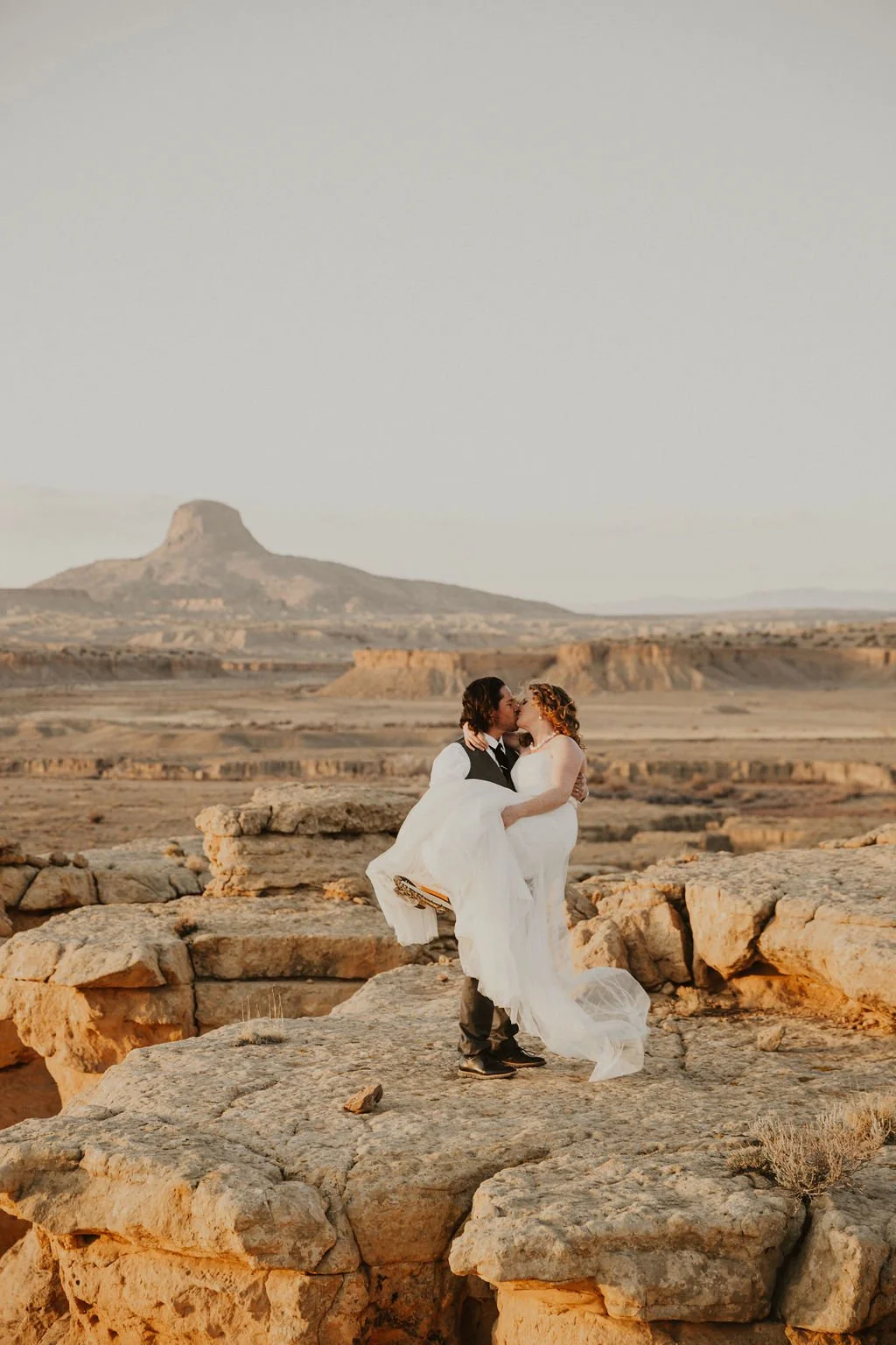 A bride and groom kissing in a desert landscape, with rocky terrain and a mesa in the background during sunset.
