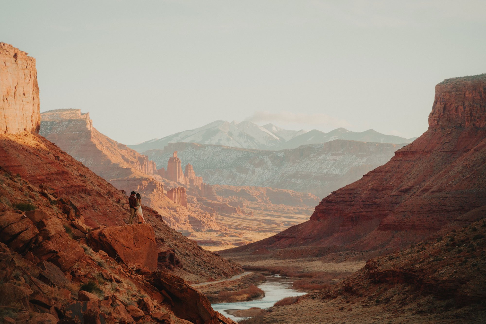 Couple standing on a red rock in a canyon landscape with distant mountains and a river.