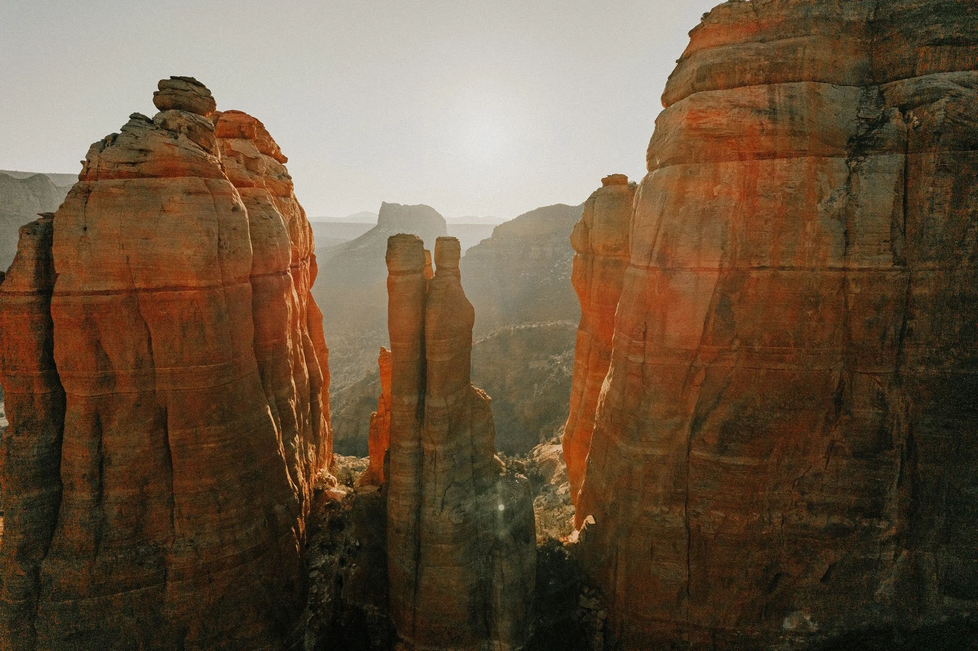 Sunlit view of tall red rock formations and cliffs in a canyon in Arizona, with a mountainous landscape in the background.
