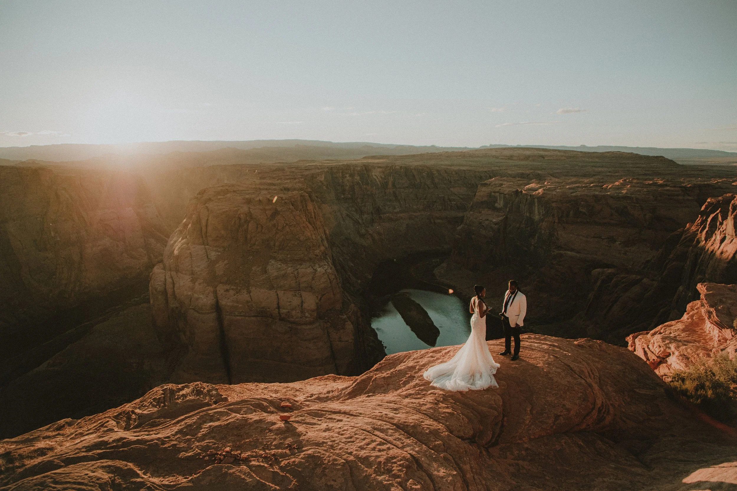 A bride and groom standing on a rocky ledge overlooking a canyon at sunset, with the river winding below them.