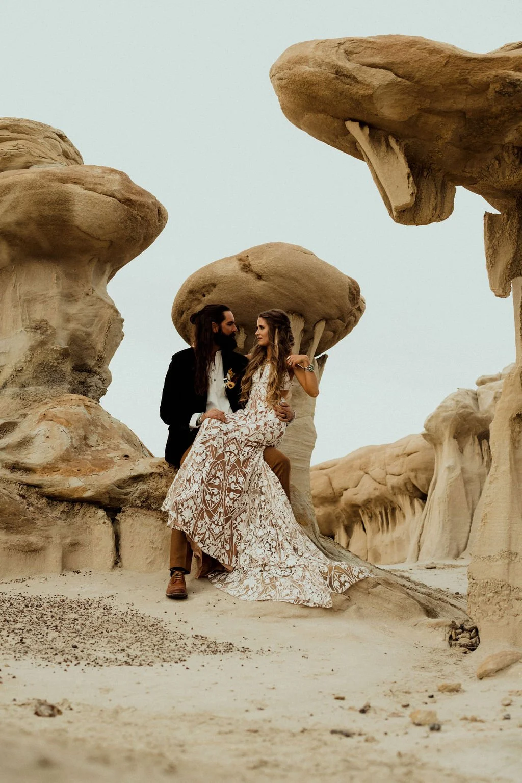 A man in a suit and a woman in a white patterned dress sit together amidst large rock formations in a desert landscape in New Mexico.