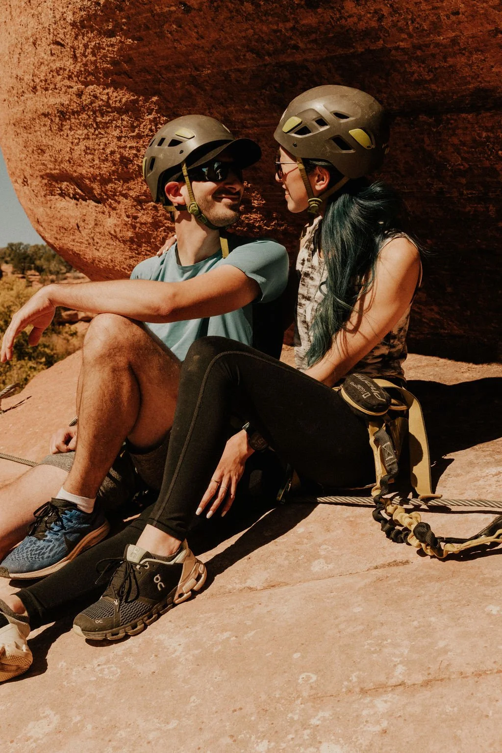 A smiling man and woman sitting on a rocky surface outdoors with large red rock behind them. They are wearing helmets, sunglasses, and hiking gear, indicating they are engaged in outdoor adventure or rock climbing.