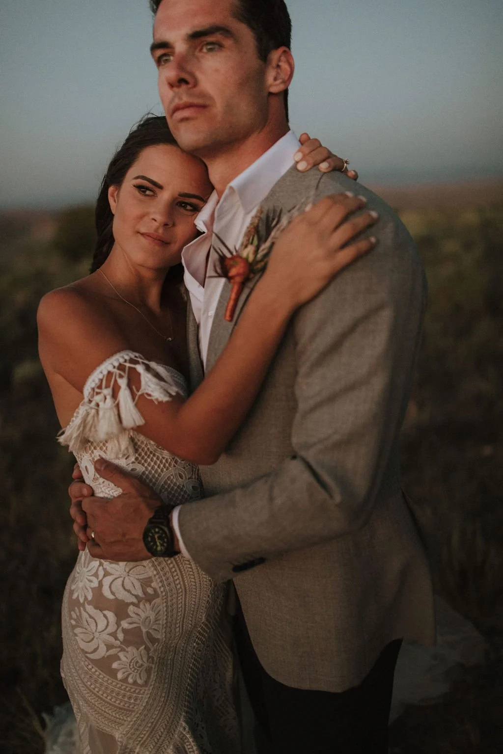 A happy couple embracing outdoors at sunset, dark sky in the background. The woman is wearing a white lace dress, and the man is dressed in a gray blazer with a white shirt. They are holding each other and smiling softly.
