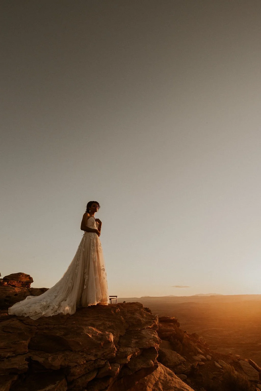 A woman in a white wedding dress stands on rocky terrain during sunset, with a vast landscape and the orange sunset sky in the background.