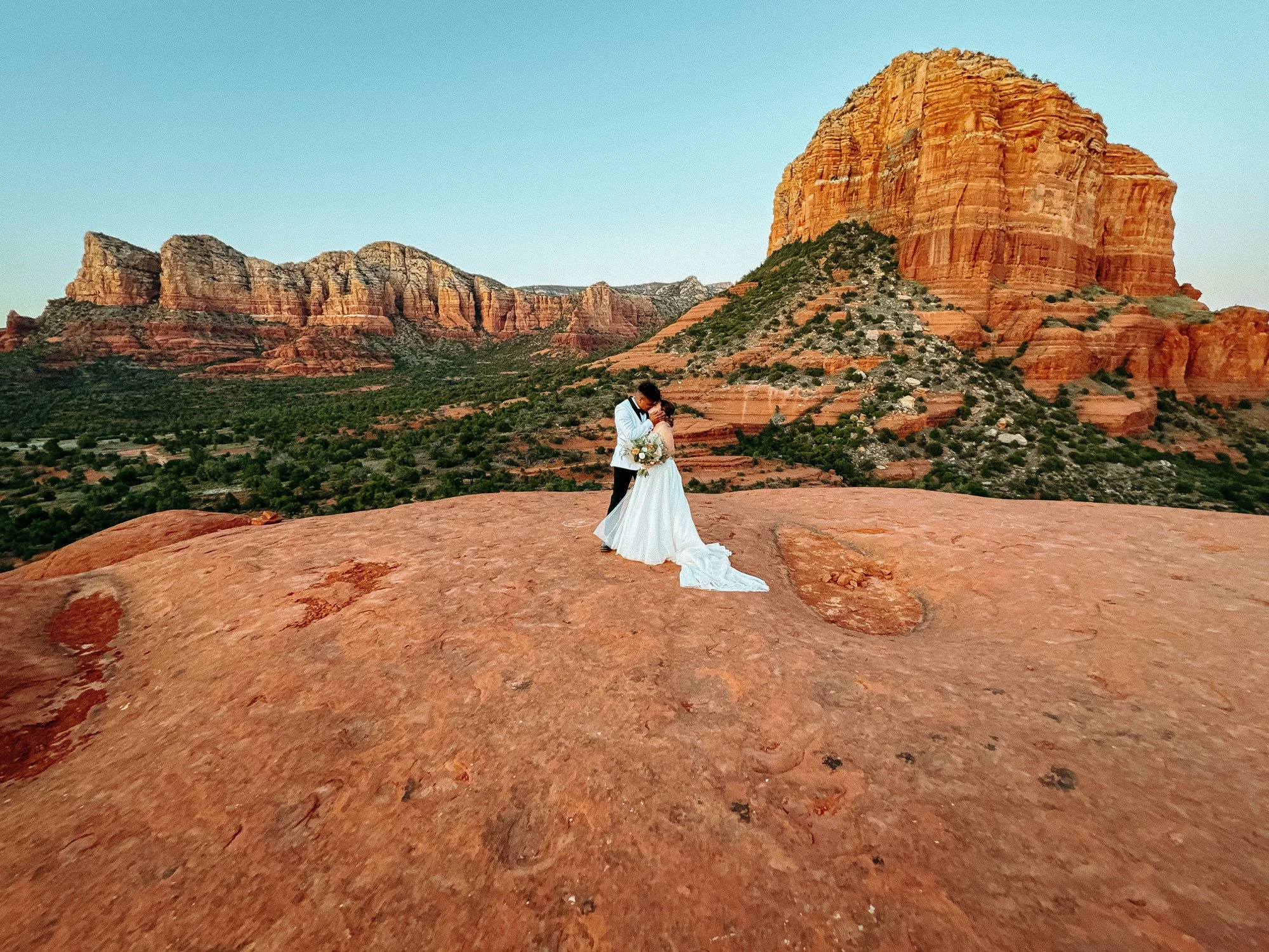A bride and groom embrace each other on a large sandstone rock in a desert landscape in Arizona with red rock formations and mountains in the background, during sunset.