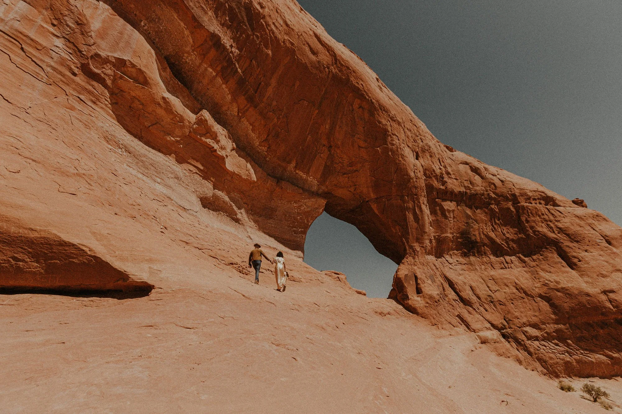 A couple holding hands walking on rocky desert terrain beneath a large natural rock arch formation, with a clear sky in the background.