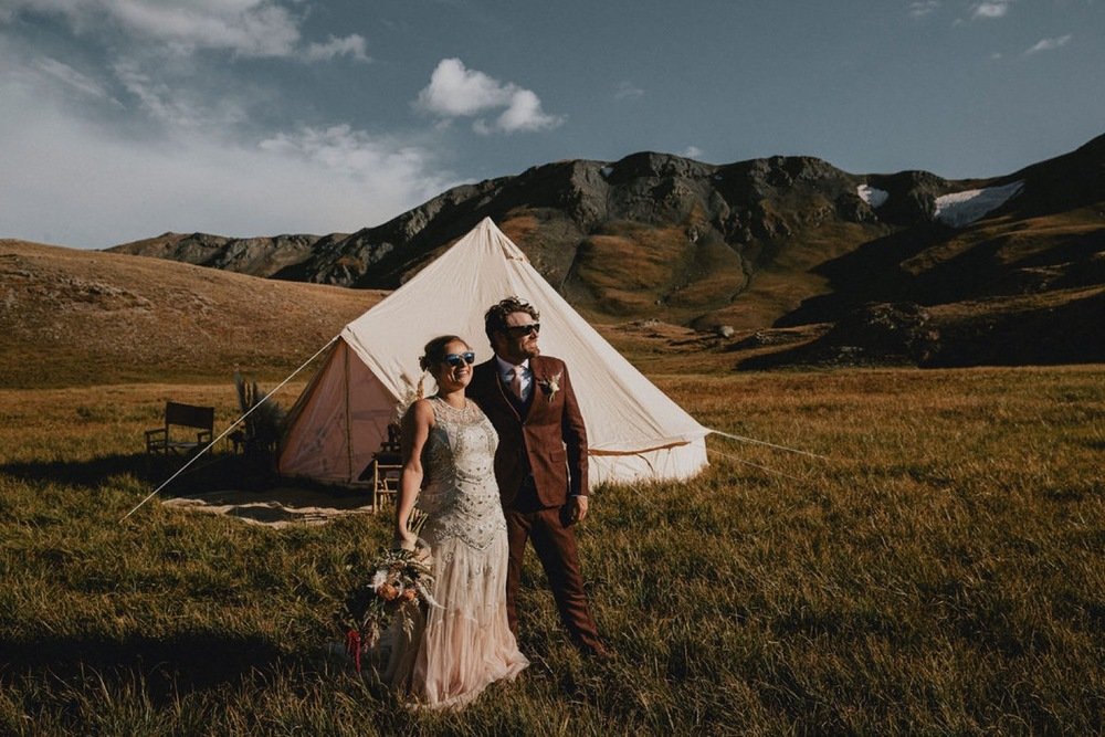 A bride and groom standing in front of a large tent in a grassy field with mountains in the background.