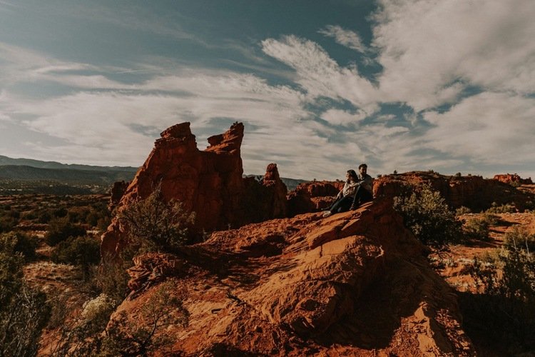 A couple sitting on a large red rock formation in a desert landscape with rock formations in the background under a partly cloudy sky.