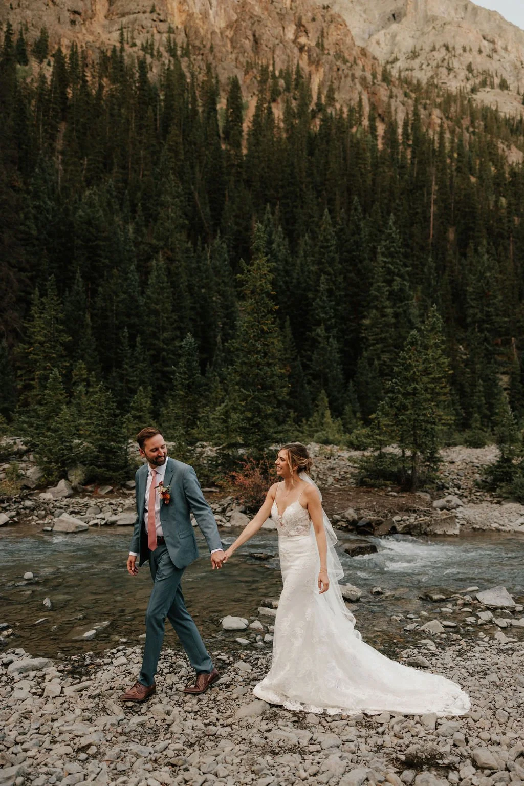 A bride and groom holding hands and walking across a rocky riverbank with tall pine trees and mountains in the background.
