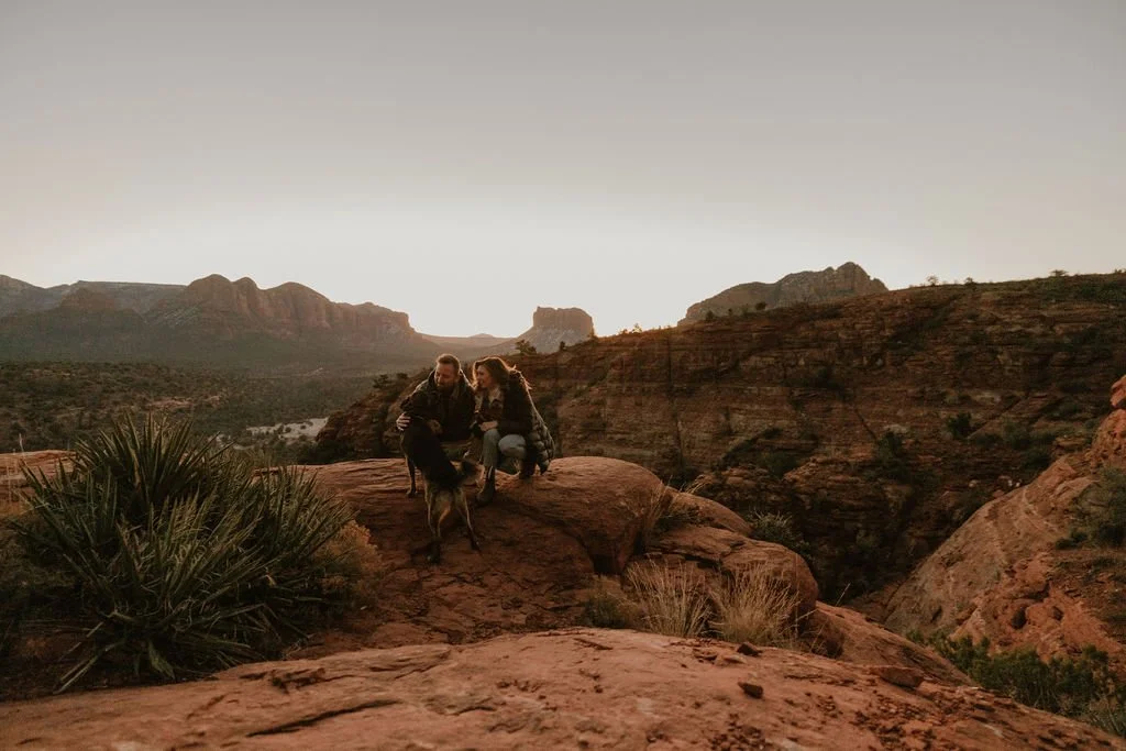 Two people and a dog on a large rock in a desert canyon at sunset, with mountainous terrain in the background.