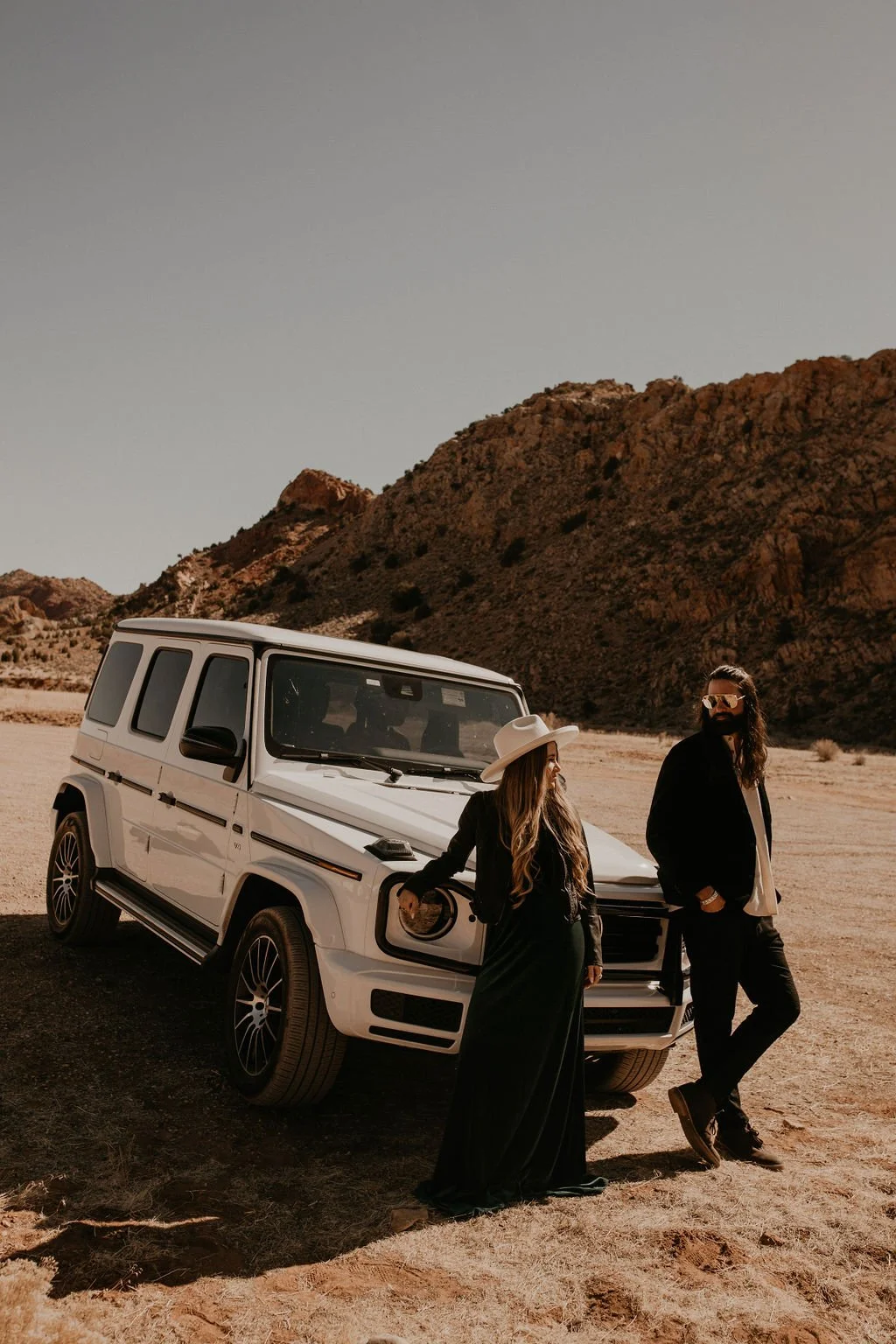 A man and woman standing beside a white Mercedes-Benz G-Class SUV in a desert landscape with rocky hills in the background. The woman wears a wide-brimmed hat and long black dress, while the man has long hair and beard, sunglasses, and casual clothin