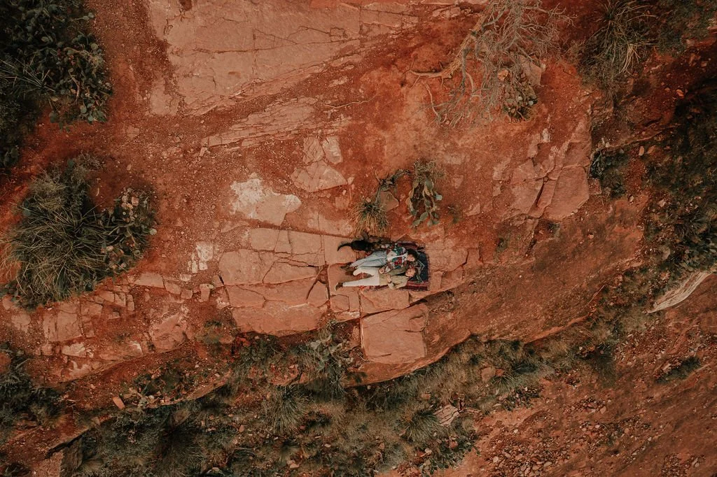 Two people lying on a rocky red-brown ground, surrounded by sparse vegetation, with an aerial view.