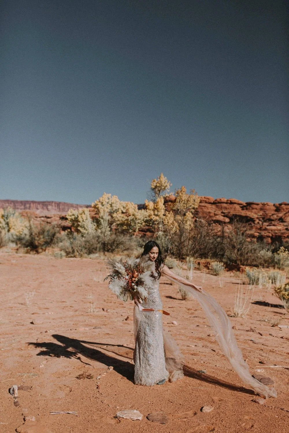 A woman in a silver dress holding a large bouquet of flowers standing in a desert landscape with sparse desert vegetation and red rocks under a clear blue sky.