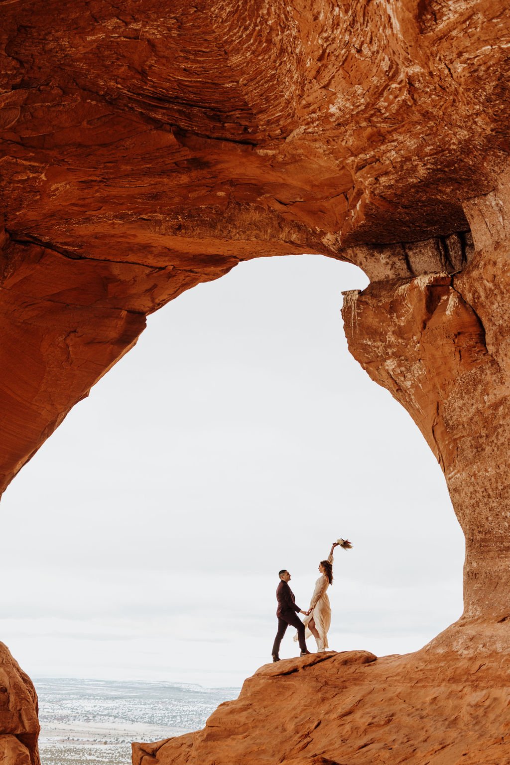 A couple holding hands and celebrating, standing on a rock between natural red rock formations, with a landscape and cloudy sky in the background.