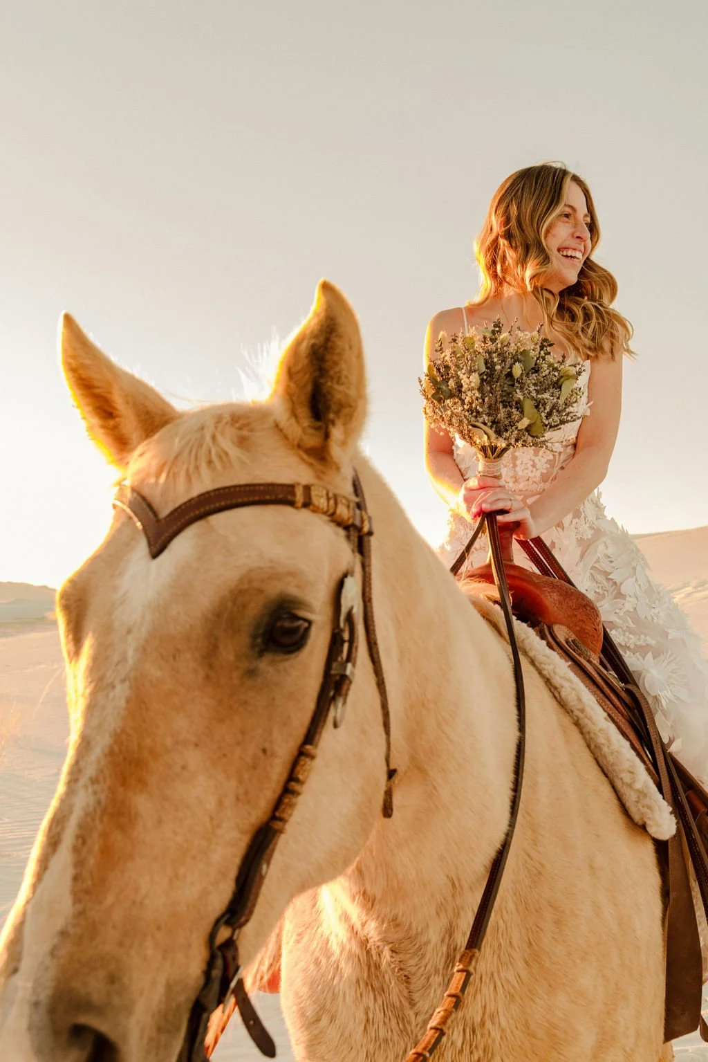 A woman in a wedding dress riding a white horse, holding a bouquet of flowers, in a desert landscape during sunset.
