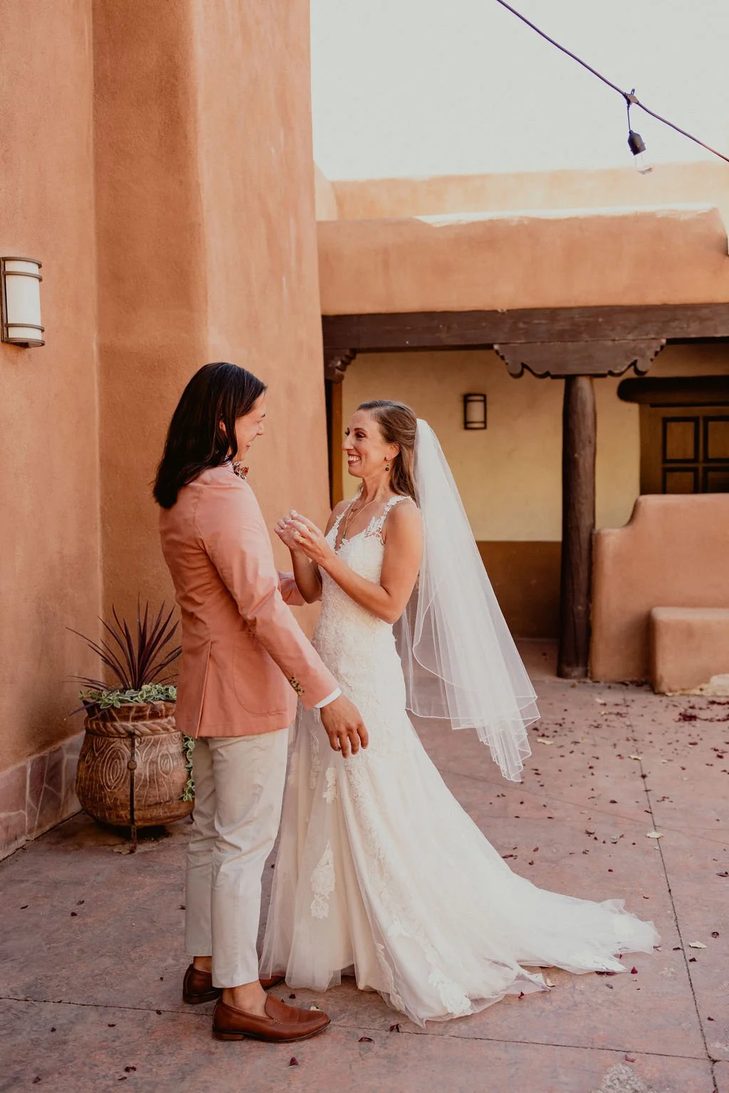 A bride and groom sharing a happy moment outside. The bride is wearing a white wedding gown and veil, while the groom is dressed in a peach suit with beige pants. They are smiling and holding hands against a rustic adobe-style building.