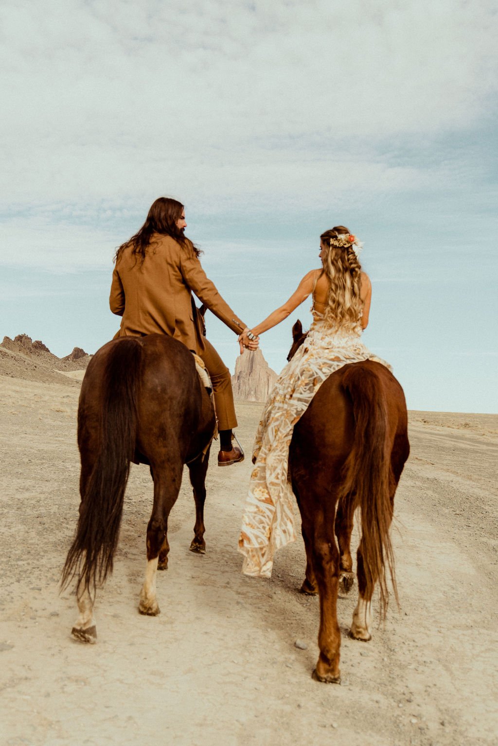 A couple dressed in wedding attire riding horses in a desert landscape, holding hands and facing each other.