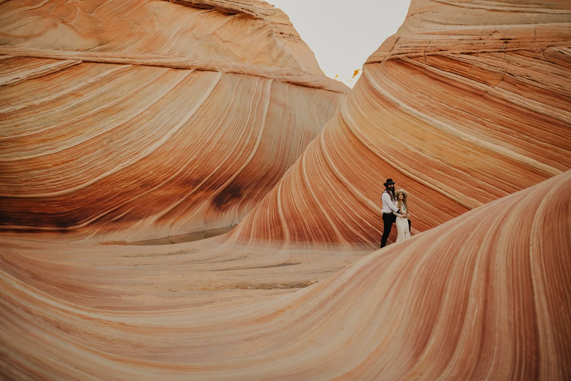 A couple stands among colorful, striped sandstone formations, likely at Antelope Canyon or a similar slot canyon, with the man wearing a white shirt and black hat, and the woman in a white dress and wide-brimmed hat.