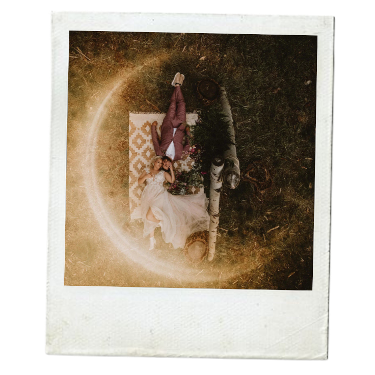 Person lying on a patterned blanket outdoors on grass, with a drink in hand and surrounded by flowers, near a fallen tree with a moon-shaped light ring around the scene.