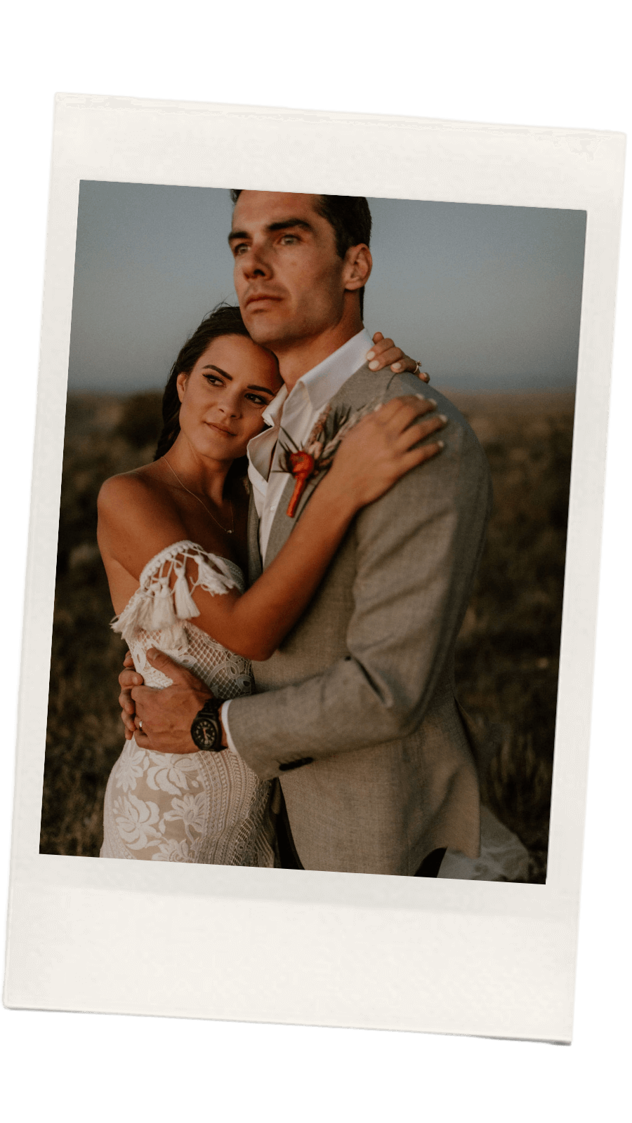 A couple hugging outdoors during sunset, the woman with dark hair wearing a white lace dress, and the man with dark hair in a gray suit with a boutonniere.