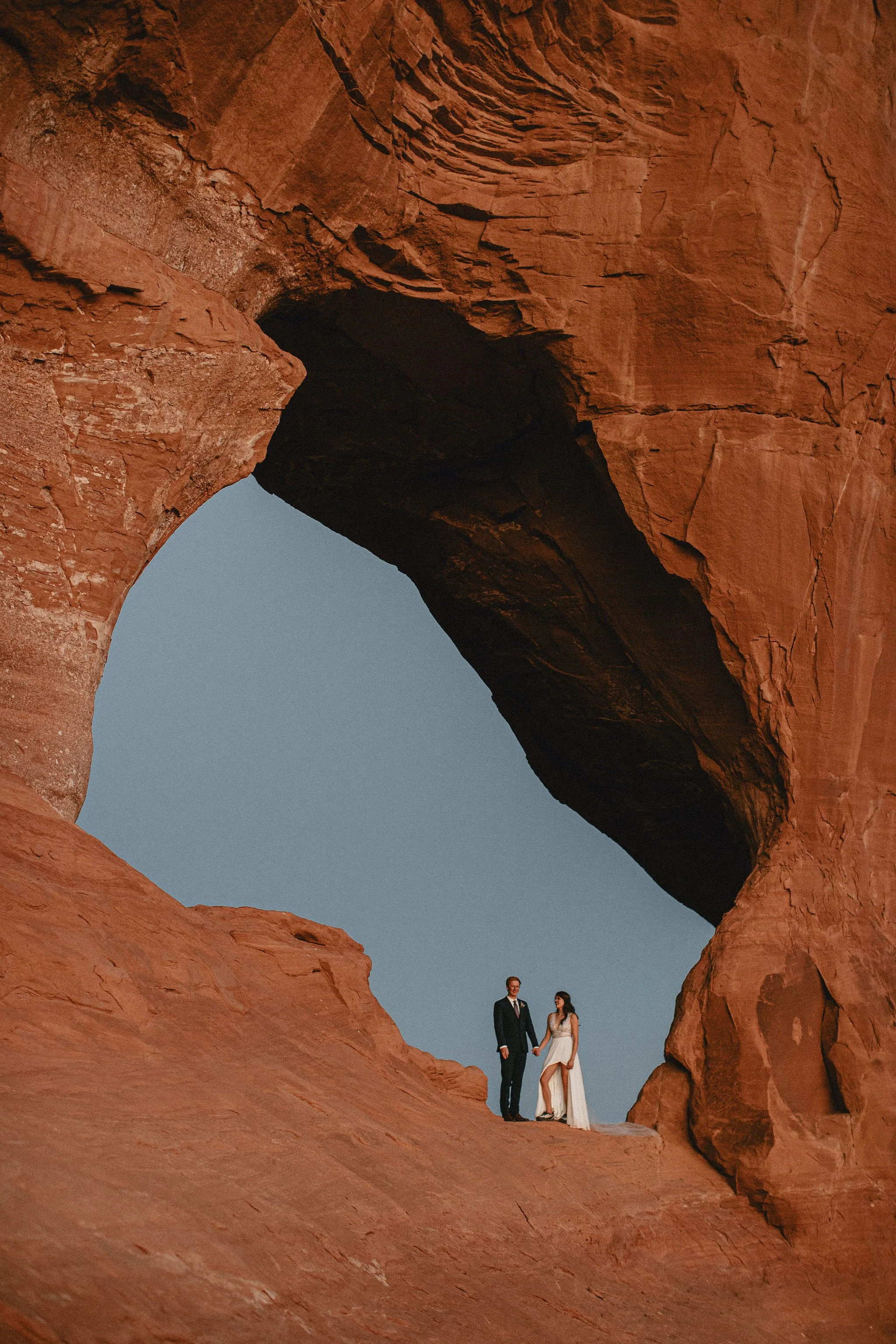 A couple in wedding attire standing inside a natural red rock arch with a clear sky in the background.
