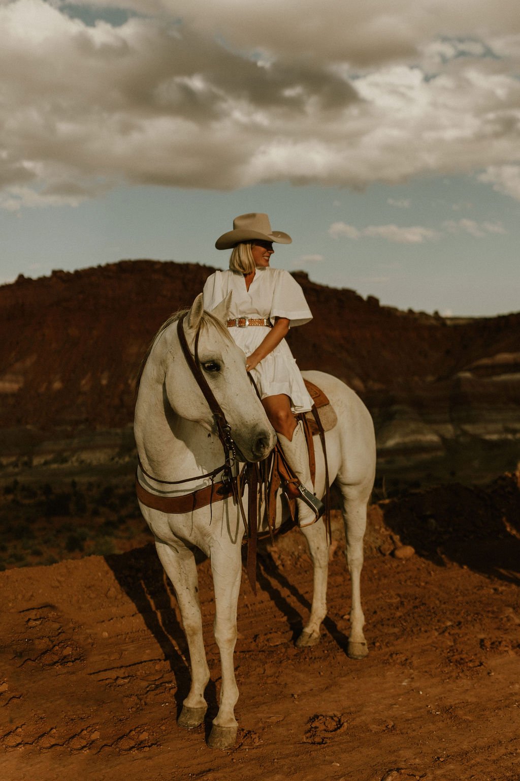Woman in a white dress and large cowboy hat riding a white horse on a dirt trail with a landscape of mesas and clouds in the background.