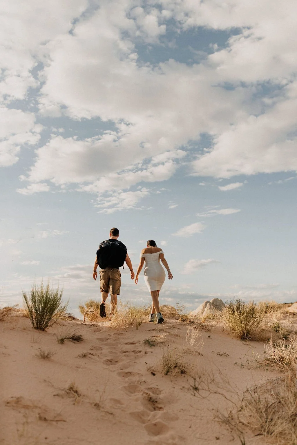 A couple walking hand in hand across a sandy desert landscape with sparse vegetation under a partly cloudy sky.