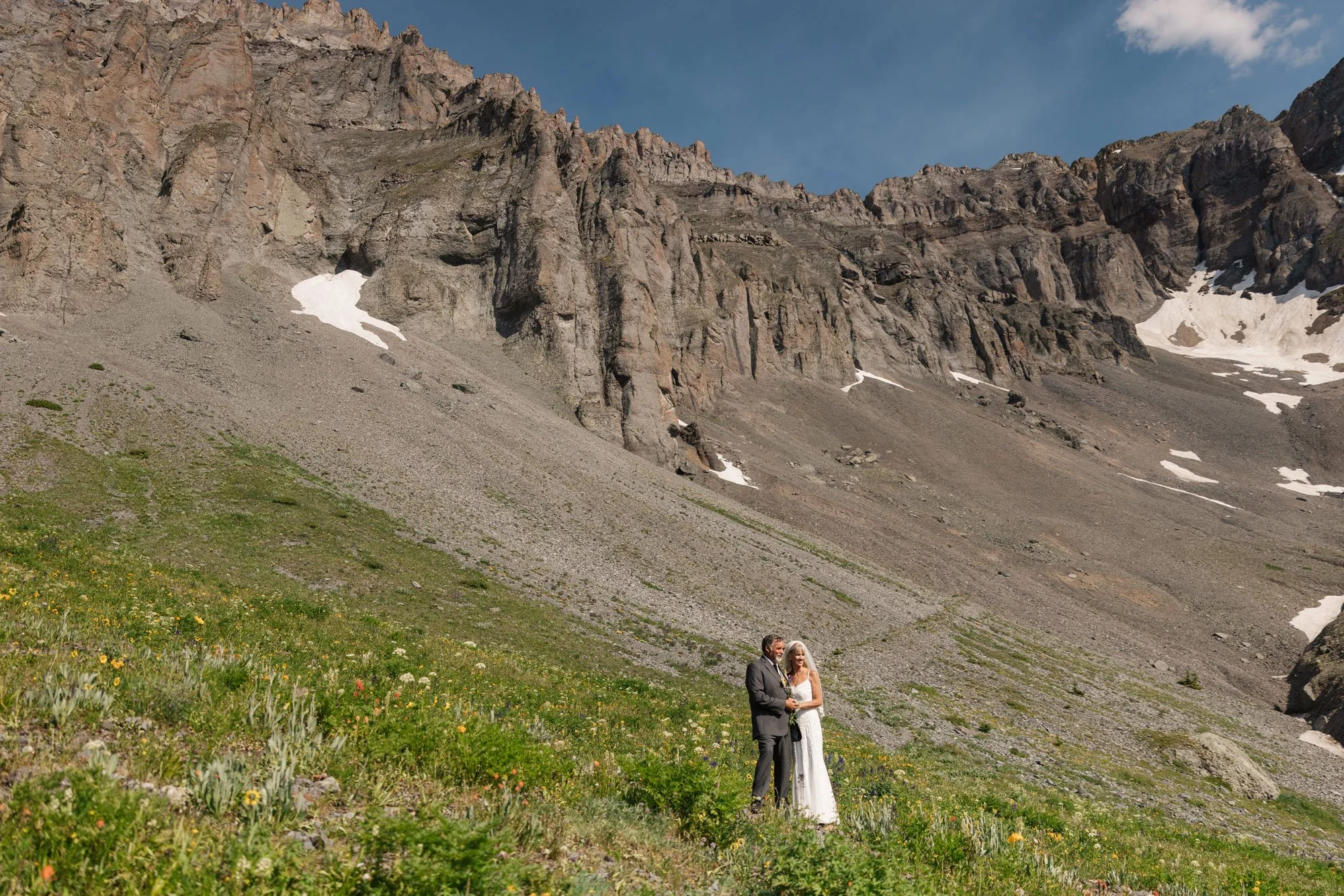A bride and groom holding hands on a grassy hillside under a mountain with patches of snow.