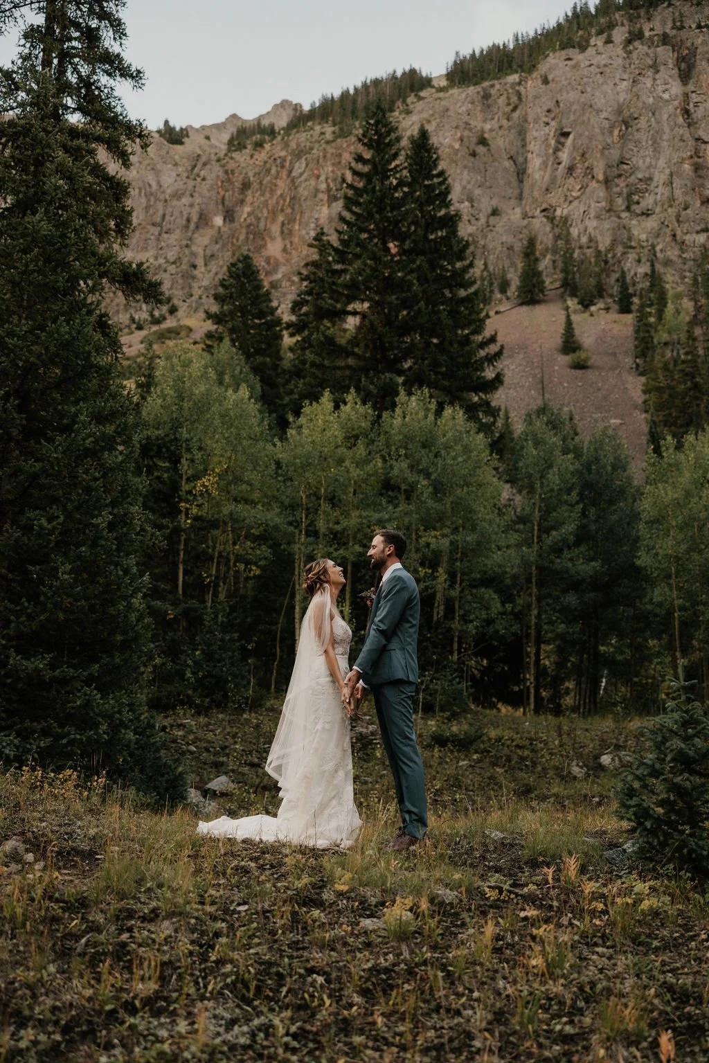 Bride and groom holding hands, standing in a forest with tall trees and mountains in the background, during a wedding ceremony.