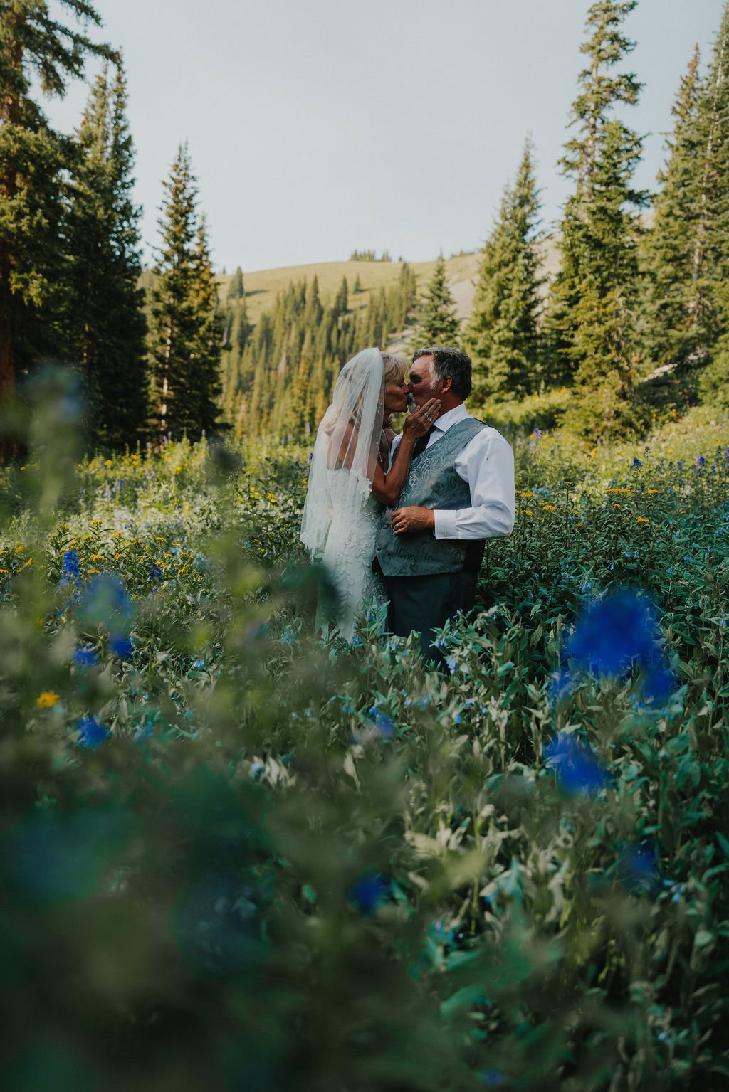 A bride and groom sharing a kiss in a field of wildflowers with trees and mountains in the background.
