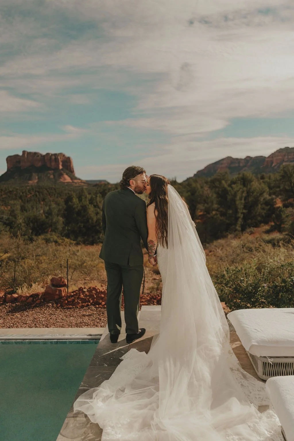 A newlywed couple sharing a kiss outdoors with a desert landscape and rock formations in the background, the bride wearing a white wedding gown and veil, and the groom in a black suit.