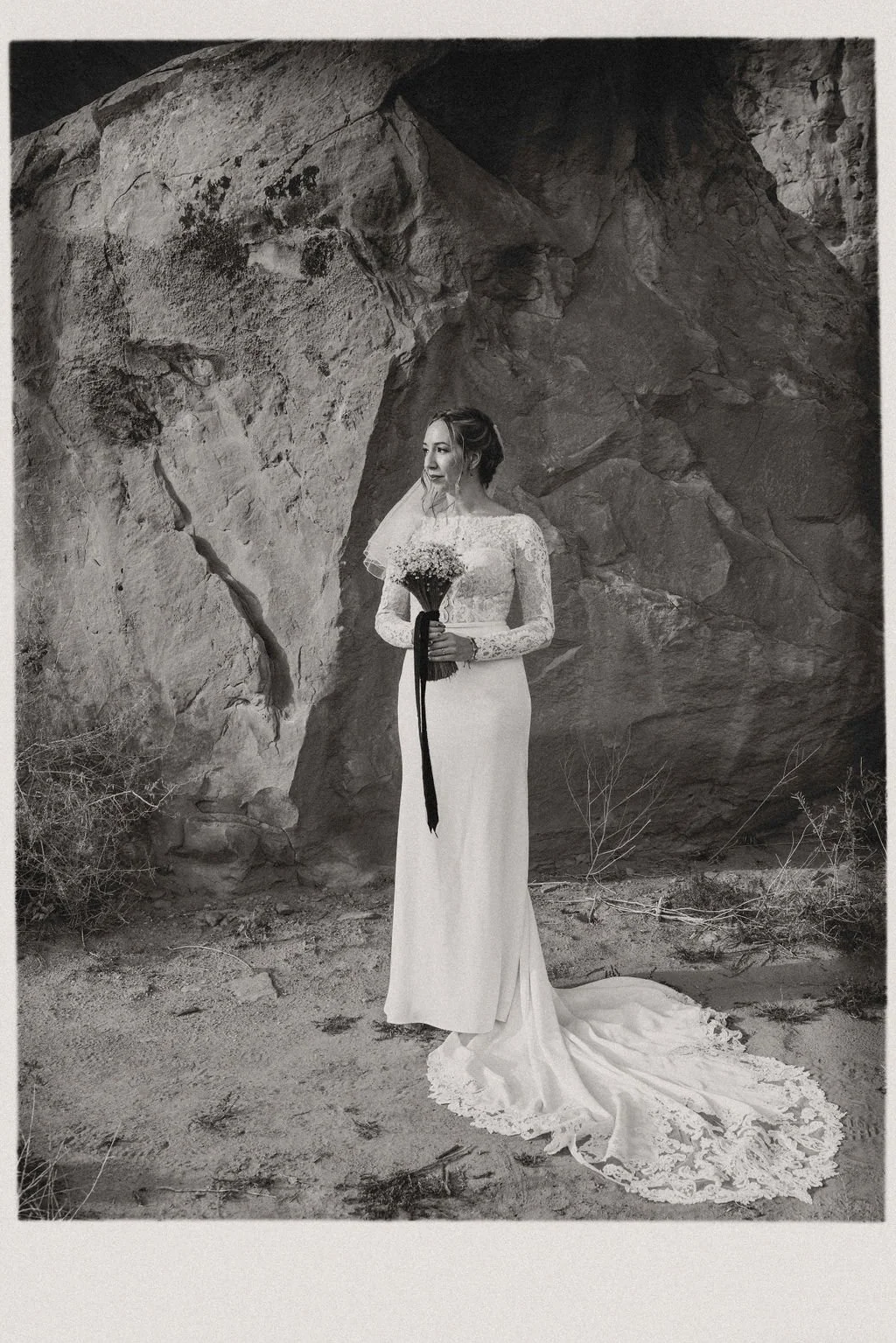 A woman in a wedding dress holding a bouquet of flowers, standing outdoors in front of a large rocky formation.