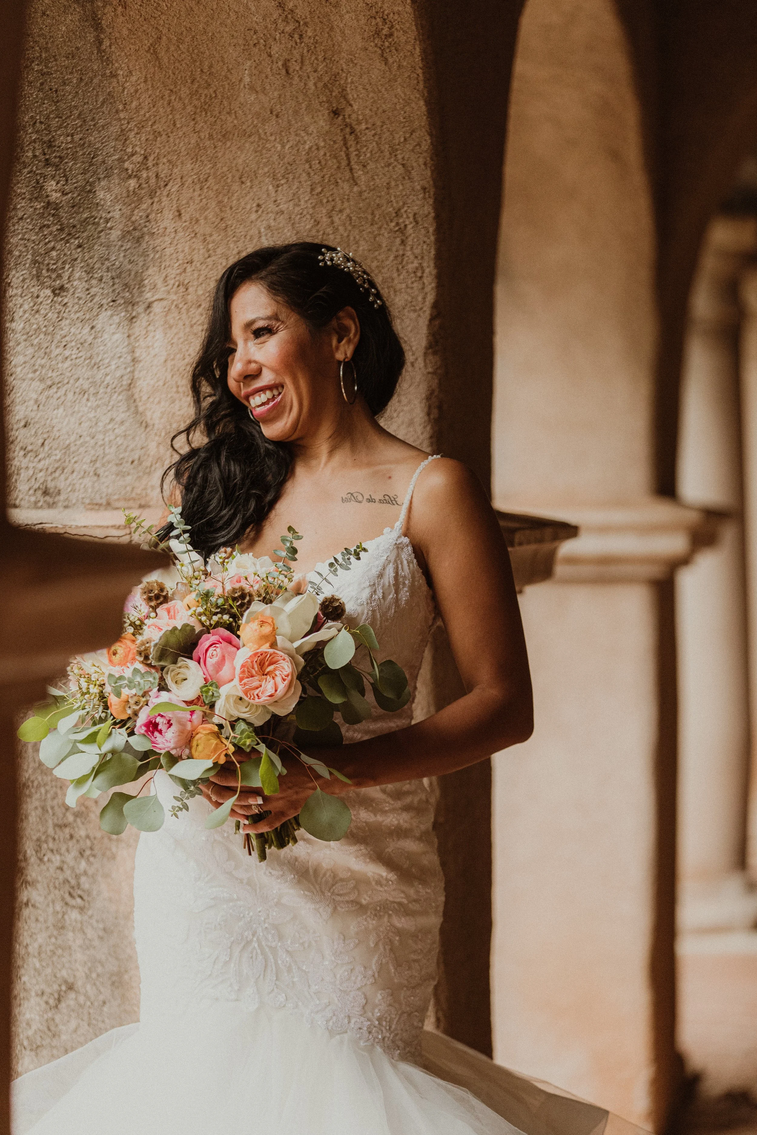 A bride in a white wedding dress holding a bouquet of pink, peach, and white roses with greenery, smiling and leaning against a textured beige wall.