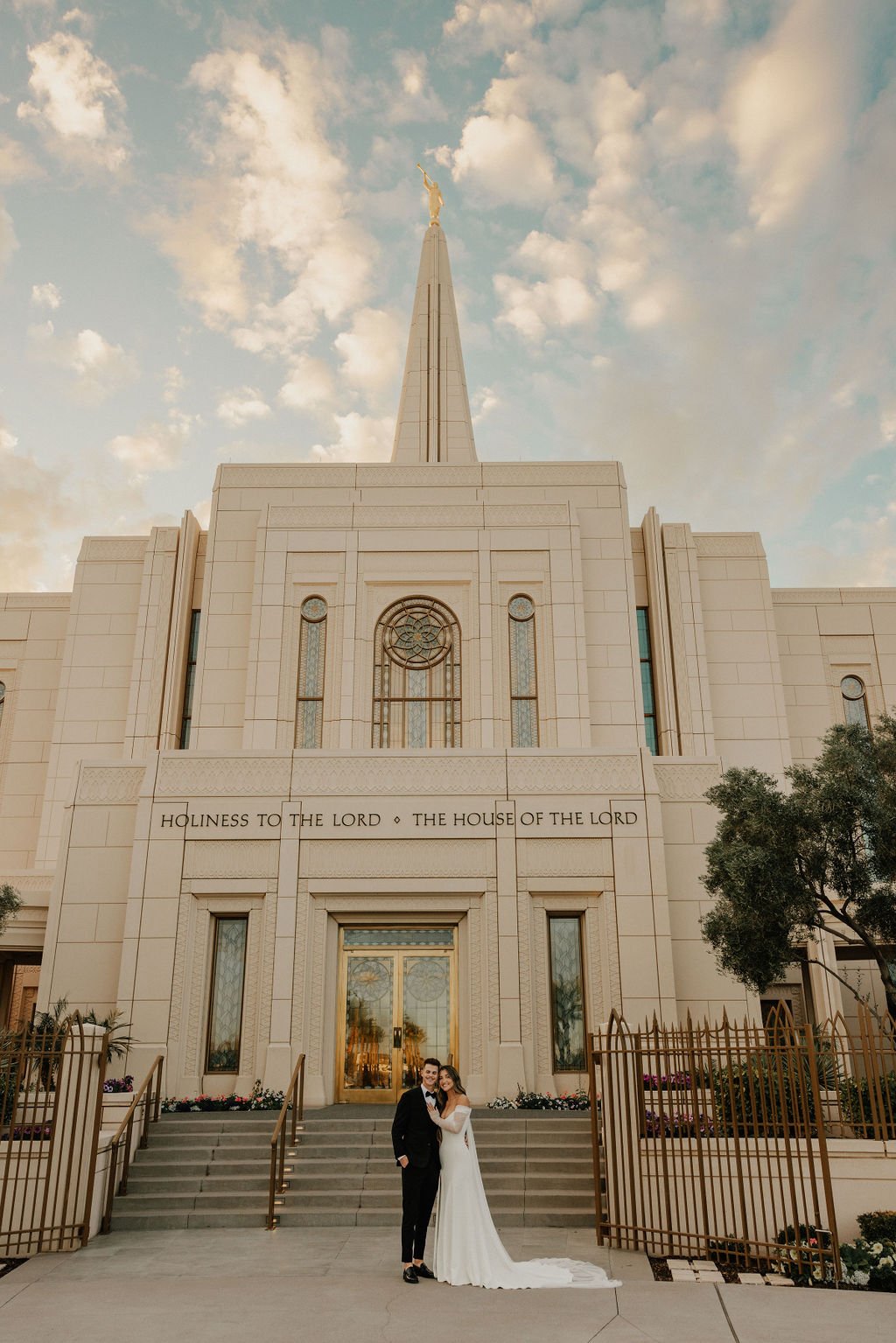 A newly married couple in wedding attire standing in front of a large church building with a steeple and stained glass windows. 