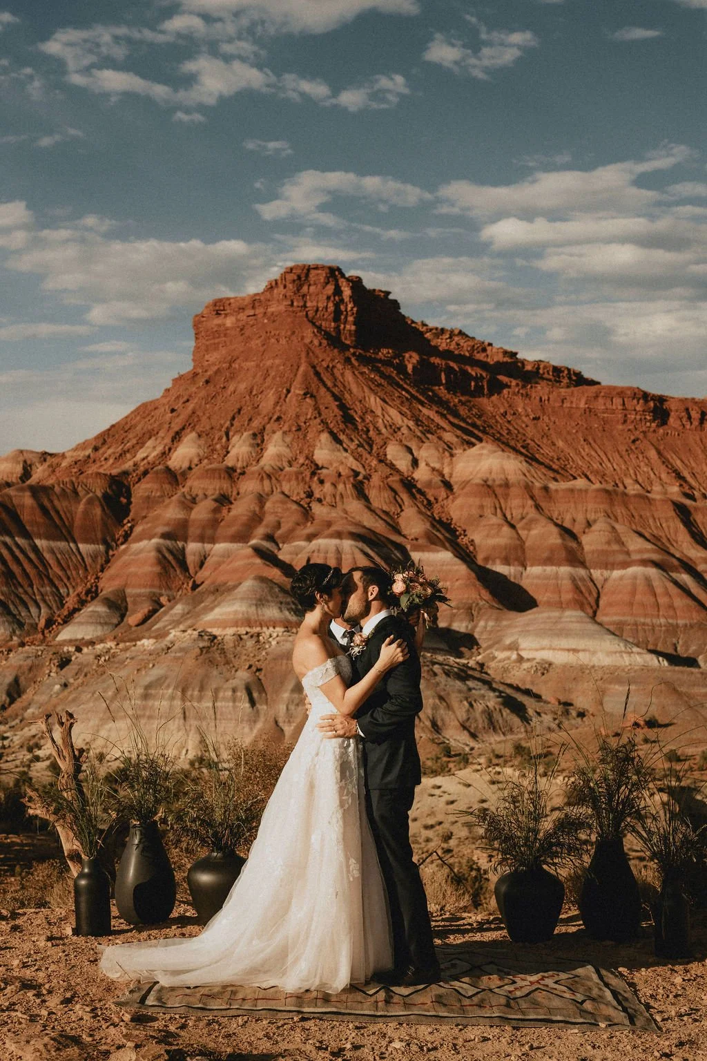 a bride and groom kissing in a desert landscape with layered colorful rock formations and a large butte in the background, in a wedding scene with vases and plants around them.