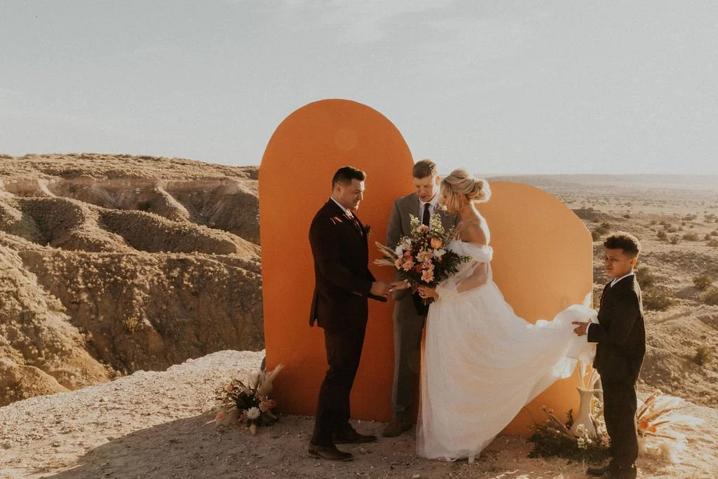 A wedding ceremony taking place outdoors in a desert landscape with a couple, a woman in a white wedding dress and two men in suits, exchanging vows in front of a large orange heart-shaped backdrop, with flowers and decorations around.