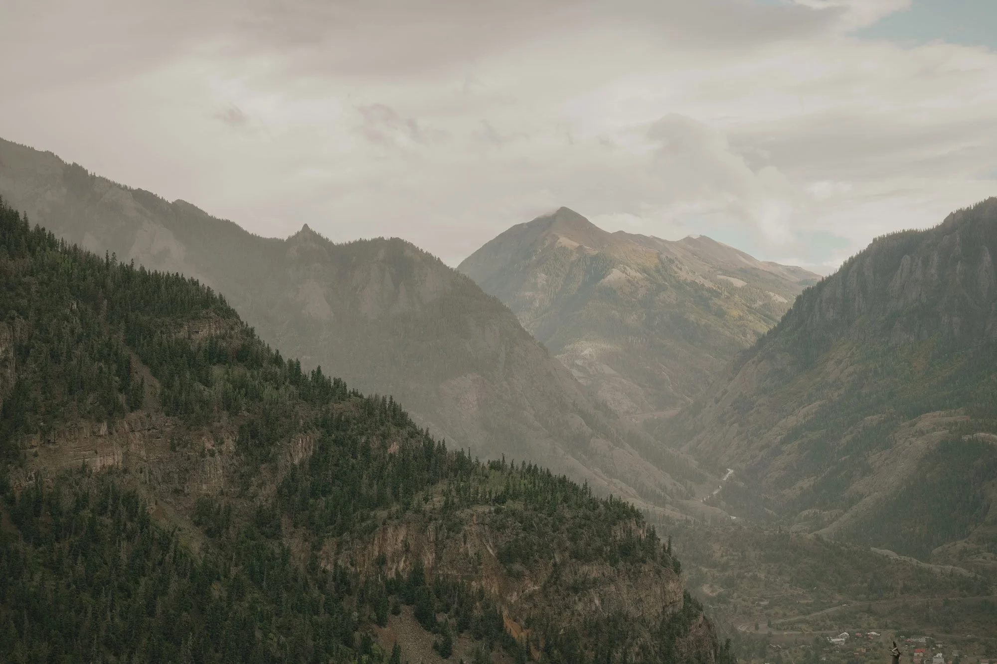A scenic view of a mountain valley with forested slopes, layered mountain ridges, and a cloudy sky in Colorado