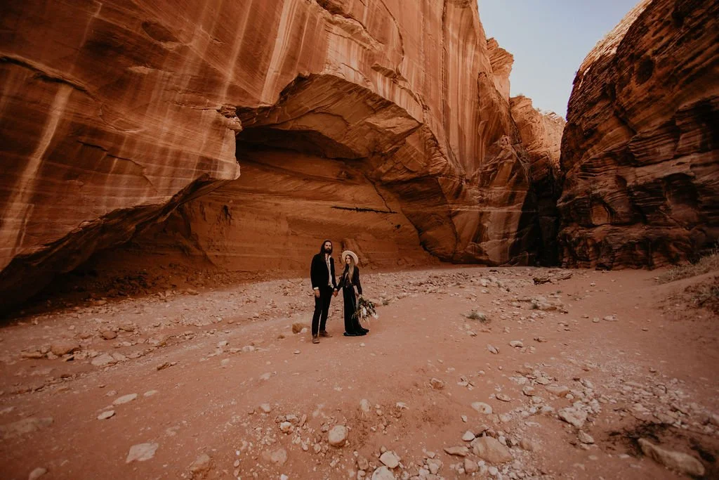 A couple standing in a narrow canyon with towering red and brown layered rock walls, desert terrain, and clear sky above.